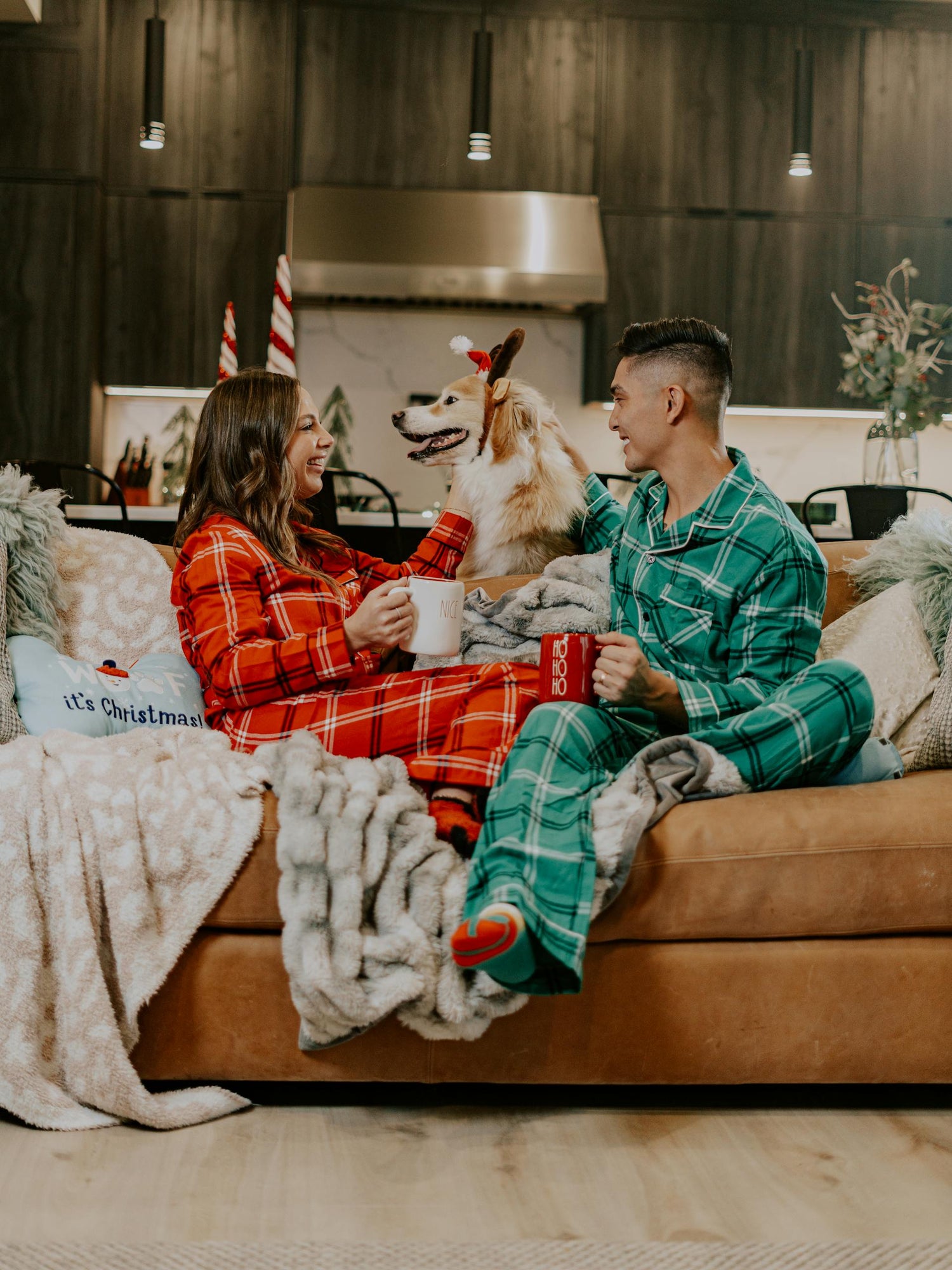 Couple wearing green and red matching winter / Christmas pajamas while sitting on a couch with their golden retriever dressed as a reindeer.  